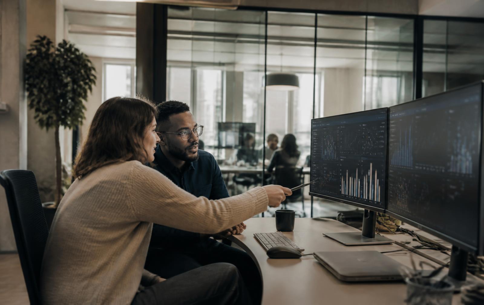 Two colleagues reviewing analytics dashboards on large monitors in a modern technology office